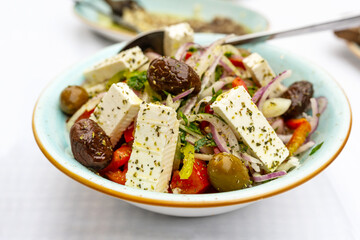 Greek salad with onions, peppers, olives, feta and herbs in a bowl, Mediterranean vegetarian dish served on a white table, selected focus