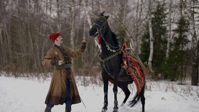 horseman in historical medieval gown and black horse in winter forest, man training stallion