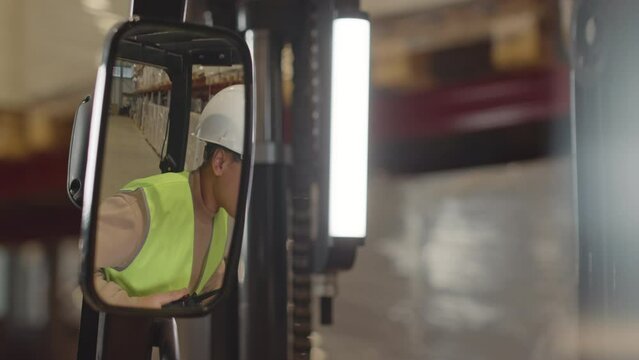 Through rear view forklift mirror shot of young Biracial female operator wearing safety hard hat and acid green vest giving instructions to employees while working together at warehouse