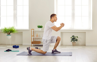 Side view of fit young man in shorts and T shirt doing forward lunge exercise on yoga mat during active fitness workout at home. Sport, wellness, physical activity, healthy lifestyle concept - obrazy, fototapety, plakaty