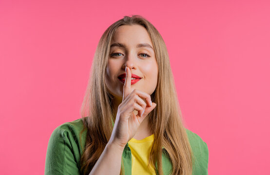 Smiling woman with finger on lips - shhh, secret, silence,pink studio background