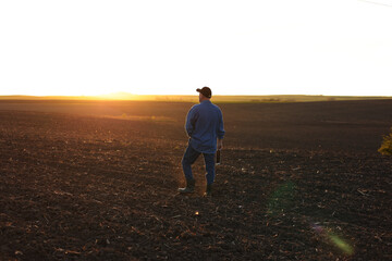 Agribusiness. Back view of male farmer walking through on cultivated plowed field at sunset in...