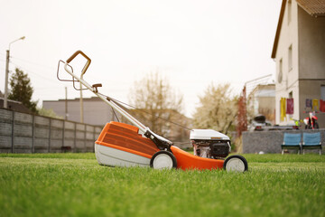 Modern orange-grey electric lawn mower on bright lush green lawn at residential backyard of house. Gardening work tools. Rotary lawn mower machine cut grass. Professional lawn care service.