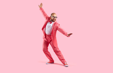 Happy handsome guy in a pink party suit dancing isolated on a pink background. Funny confident bald young man wearing a suit, bowtie, sneakers and glasses dancing and having fun in the studio