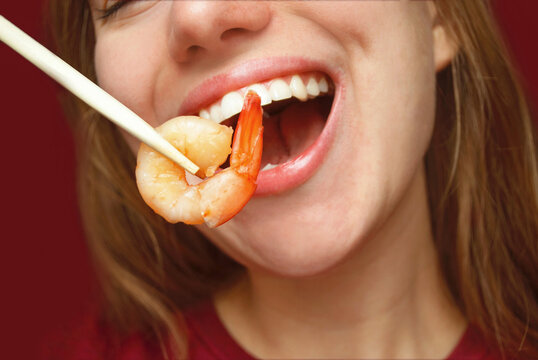 Happy woman eats big appetizing shrimp with chopsticks. Close-up portrait, red background.