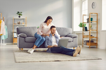 Young married couple sitting on sofa in the living room at home using tablet PC for internet and social media. Happy family resting on couch enjoying weekend watching video or talking on video call. - obrazy, fototapety, plakaty