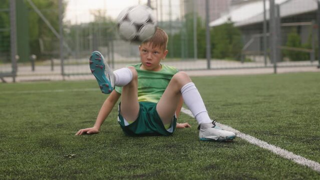 The boy dressed in football uniform is practicing on a mini football stadium. A portrait of a young football player is attempting to kick the ball while lying down.