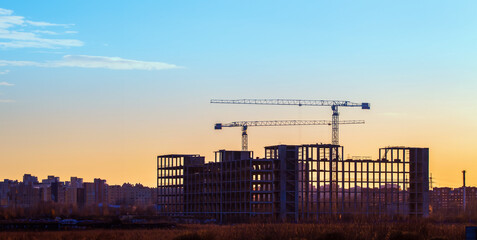 Fototapeta na wymiar Construction landscape. Tower cranes at sunset. Frame of multi-storey building under construction. Panorama with unfinished houses. Construction panel buildings. Building landscape with summer sunset