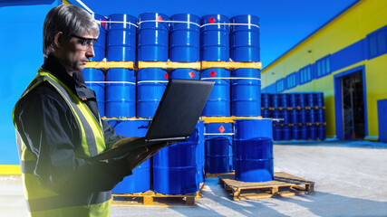Warehouse worker. Man with laptop. Guy near blue barrels. Warehouse of chemical enterprise. Chemical storage worker. Petrochemical warehouse manager. Pallets with barrels near industrial building