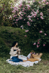 young beautiful brunette woman sits on a picnic blanket near a lilac in the park. woman drinking coffee or tea in cermic cup outdoors. picnic breakfast	