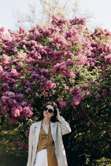 a young beautiful woman in linen clothes in a casual style stands in a lilac garden. a woman in sun glasses stands near a blooming lilac.	