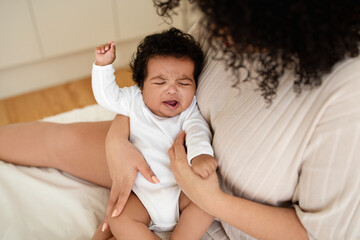 Black millennial mom calms crying sad little baby on white bed in bedroom. Disease treatment, teeth...