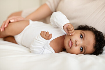 Fototapeta na wymiar Glad cute black little baby in clothes lies on white bed with mom hands in comfort bedroom, close up