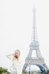 Fototapeta na wymiar Lady with flowing blond hair, in white clothes softly touching face with hand on background of famous Eiffel Tower.
