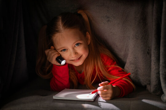 Cute Smiling Girl Hiding Under Blanket With Flashlight And Writing In Notebook