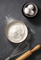 Natural organic flour in a bowl with a eggs and rolling pin on a dark gray background. Ingredients for home baking.