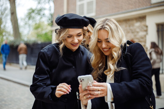 Two stunning friends smiling while watching photos on phone. Blonde with flowing hair show screen to woman with beret.