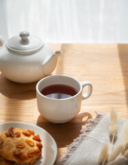 A cup of tea with homemade cookies and a white teapot on a wooden table against the background of a window with sunny morning light and shadow.