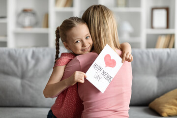 Cute Little Girl Holding Handmade Greeting Card And Embracing Mom At Home