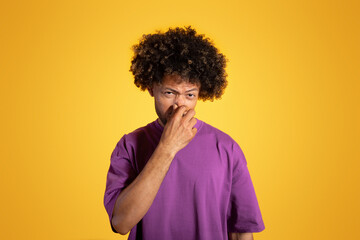 Fototapeta na wymiar Despaired sad mature black curly man in purple t-shirt covers nose with hand, suffers from bad smell