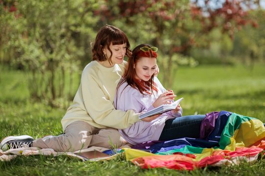 Picnic time in the middle of nature lesbian couple very in love in the park sitting down on the blanket and reading together a book and smiling large - Powered by Adobe