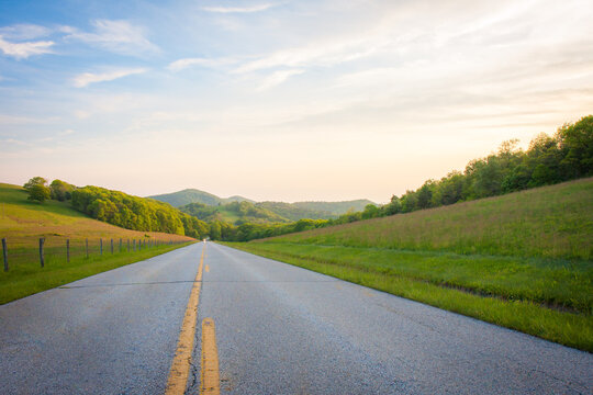 Paved road through rural grassy fields and mountain ranges at sunset