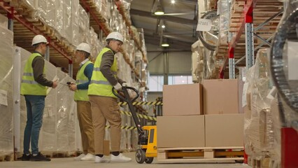 Full length shot of female warehouse worker carrying hand pallet truck loaded with parcels packed in cardboard boxes along aisle with other goods prepared for dispatching - Powered by Adobe