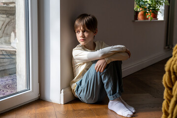 Kid psychological trauma. Lonely child boy sadly look to window hug knees sits on floor at home...