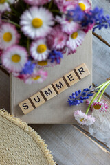 Bouquet of wild daisy flowers in a cup, straw hat, book. Cozy still life on the window of an old countryside house, summer cottage. Floral home decoration. Word summer written in wooden letters
