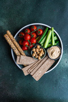 Overhead view of a plate of hummus, chickpeas, crispbread, breadsticks, tomatoes and cucumber batons