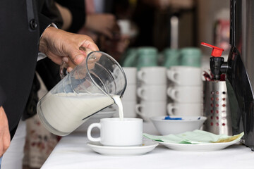 Frau füllt am Buffet Milch aus einer Karaffe in eine Kaffeetasse - obrazy, fototapety, plakaty