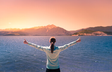 Young woman facing mountain lake sunset rejoices, smiles looking up to the sky, enjoys life and...