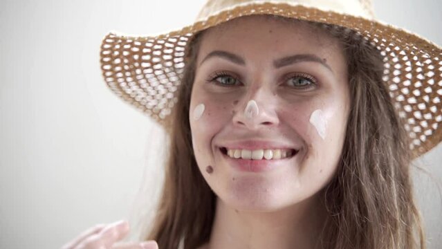 A young woman in a straw hat puts on sunscreen. She has many moles on her face