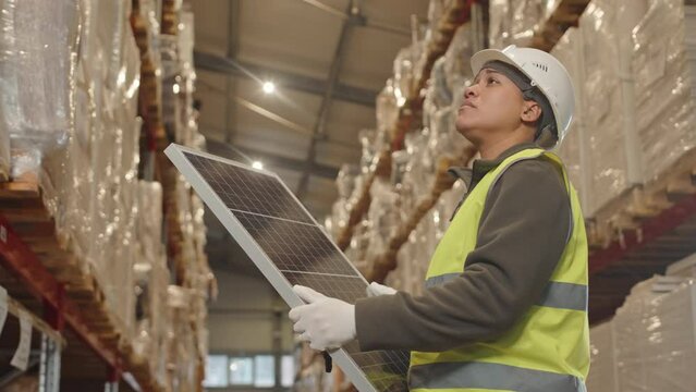 Low angle medium shot of young Biracial female warehouse worker wearing hard hat and acid green vest using solar battery at workplace