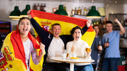 Company of excited young adult sports fans waving flag of Spain and supporting national team with...
