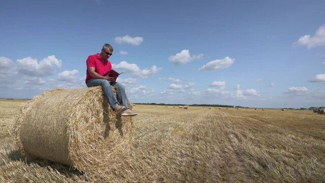 adult man in sunglasses sits on straw bale on beveled field and makes recordings in tablet