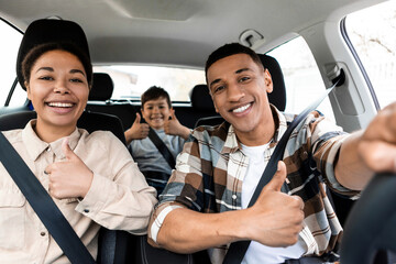 Happy african american family of three driving new car and gesturing thumbs up, approving automobile, like this auto and smiling