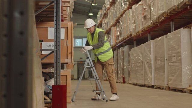 Full length shot of Biracial female warehouse worker in safety uniform climbing up ladder to catch for parcel in cardboard box on shelf