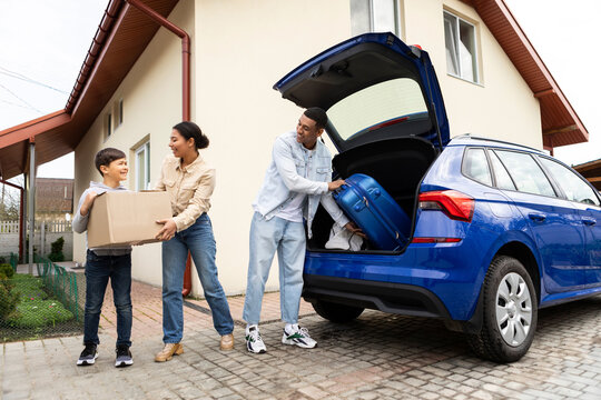 Happy african american family preparing for holiday, putting suitcases and boxes in car trunk, ready to go on vacation trip, child boy helping parents