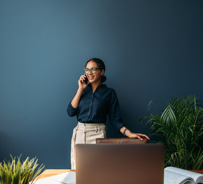 Smiling entrepreneur in formal wear talking on a cell phone while standing at a blue wall at home