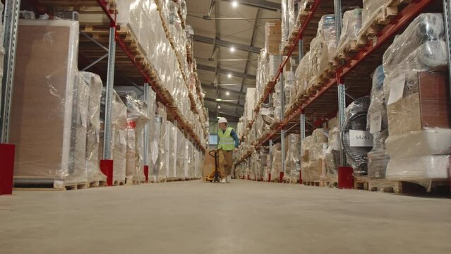 Low angle stab shot of mature male warehouse worker in uniform carrying hand pallet truck with cardboard boxes on it along big warehouse