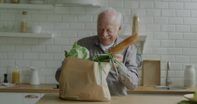 Elderly man carrying shopping bag then unpacking organic food in kitchen in apartment. Grocery products and retired person lifestyle concept.