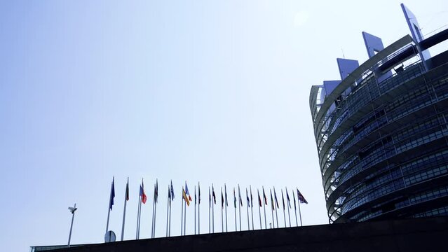 Side view of all european union country members flag waving in near glass building with surveillance camera on the nearest pole surveilling the zone