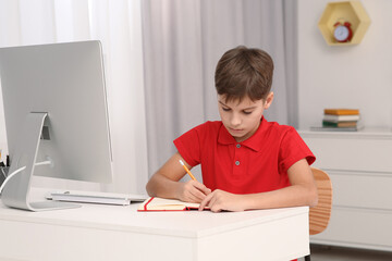 Boy writing in notepad while using computer at desk in room. Home workplace