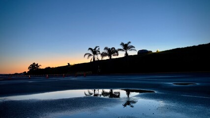 Wide view, silhouetted SUV and palm trees at top of hill with orange sunset sky in background and reflections on puddles in parking lot in foreground