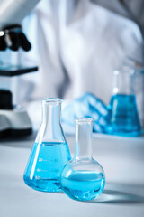 Closeup view of scientist pouring light blue liquid from test tube into flask and laboratory glassware on table, selective focus