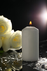 White roses and burning candle on black mirror surface in darkness, closeup. Funeral symbols - obrazy, fototapety, plakaty