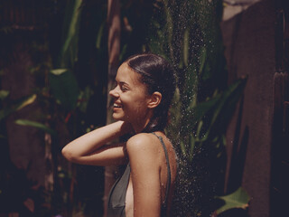 Fototapeta na wymiar A woman, a body in a swimsuit washes her head in a tropical shower outdoors against backdrop green tropical leaves, flowers and palm trees. Body and hair care, tanned skin, sunlight, smile, vintage
