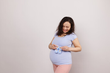 Beautiful smiling pregnant woman in blue t-shirt and pink pants, holding blue knitted baby booties over her belly, isolated white background. Gener reveal party. Newborn clothing. Pregnancy. Maternity