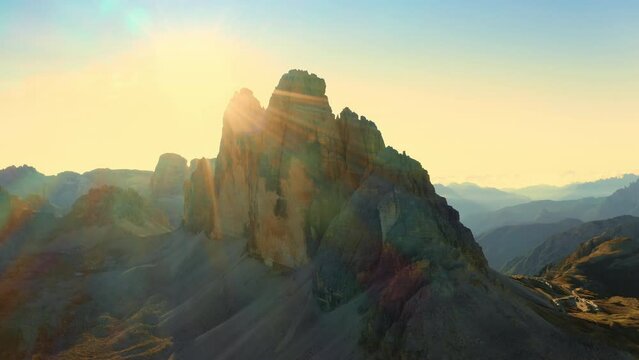 Sunrise illuminates mountainous terrain, showcasing majestic Three Peaks of Lavaredo and sand-covered foothills in Alps, creating a captivating backlit aerial view.